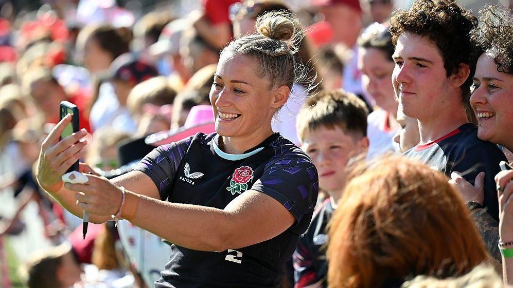 Natasha Hunt, wearing a blue England shirt, smiles while taking a selfie with a crowd of fans behind her