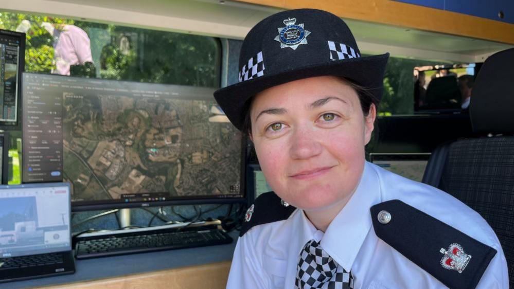 Supt Taryn Evans inside the drone control van. She is looking at the camera and there is a monitor and laptop behind her. She is wearing a small police officer's hat, a blue shirt with dark blue epaulettes, and a chequered tie.