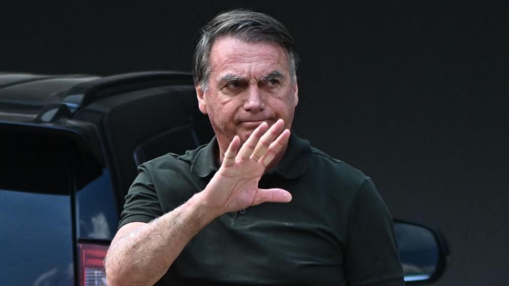 A shot of Brazil's former president Jair Bolsonaro waving to the right, dressed in a dark green shirt and hair combed to the right, while walking in front of a car.