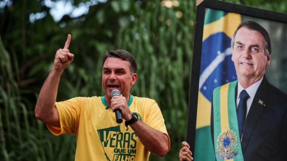 Brazilian presidential candidate Flavio Bolsonaro of the Liberal Party (PL) speaks next to a portrait of his father, Brazil's former President Jair Bolsonaro, during a campaign rally in Goiania, Brazil, September 27, 2026. He is wearing a yellow T0shirt and holds up his fight index finger as he speaks into a microphone.