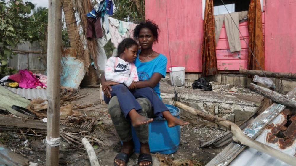 A mother and daughter sit outside their home with fallen tree branches and other debris surrounding them. The mother is wearing a blue top and the daughter a white Nike jumper.