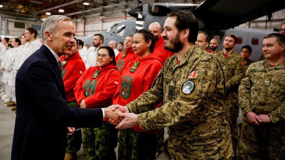 Prime Minister Mark Carney is seen shaking hands with a service member of the Canadian Armed Forces. Behind them are rows of service members from different units, standing in front of a military aircraft inside a hangar.