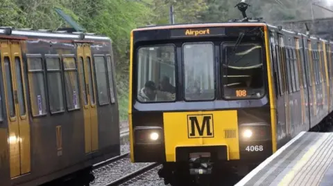 Front view of a Tyne and Wear Metro train, which is just pulling into a platform, with the destination showing as Airport. A passenger can be seen in the front seat. On the opposite rails is a side view of another metro train with two doors visible.