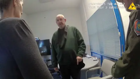 Murdoch wearing dark green jumper stands in front of camera while questioned by two officers in front of him