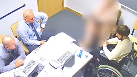 Kyle Clifford in a wheelchair in front of two police officers who are behind a desk. There is a blurred-out person next to Clifford.