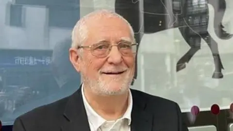 A close-up image of a man with silver hair and a light beard smiling at the camera. He is stood in front of a window with the Coventry flag in it - a blue and white flag with an image of a woman on a horse in the middle.