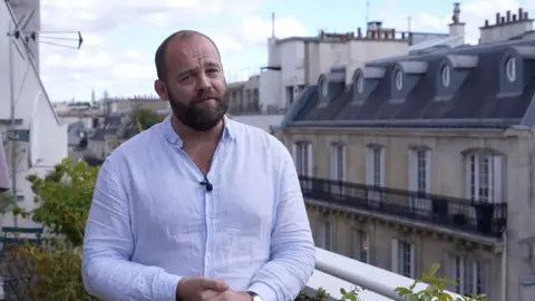 BBC Correspondent James Waterhouse wearing pale blue shirt and standing on balcony