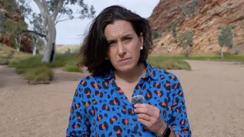 The BBC's Katy Watson wearing a blue leopard print blouse stands in front of a rocky outcrop