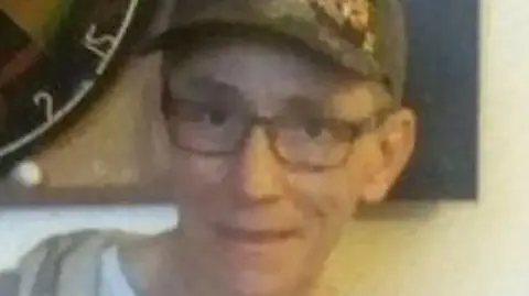 Vincent Cullinane is wearing glasses and a green baseball cap. He is smiling while pictured in front of a dart board.