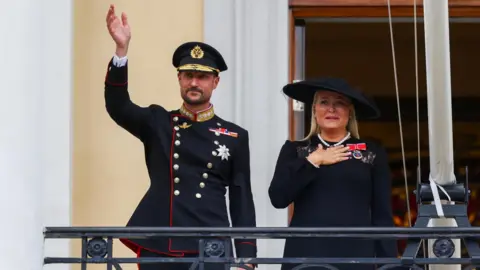 King Haakon waves to the crowd from the balcony of the royal palace, as Queen Mette-Marit stands next to him
