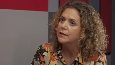 Hannah Ingram-Moore sitting down in an office room, with curly brown but highlighted blonde hair and wearing a floral top.