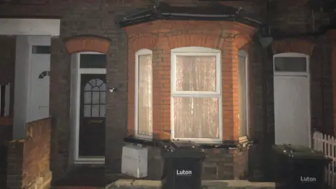 The exterior of the ground floor of a redbrick terraced house, which has a bay window and the curtains drawn, with the light on inside. The front door is to the left and outside are two wheelie bins, with the word "Luton" on the front.