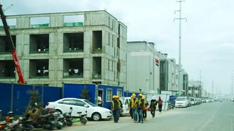 Construction workers in hard hats walk along a wide road lined with large warehouse-style data centres under construction.