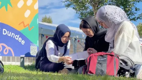 Three women sitting on grass looking at coursework together. They are near a sign that says Dundee.