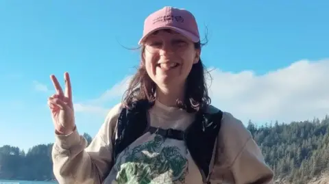 Paul Burke Becky Burke outdoors in front of a forest which can be seen in the far distance. The sun is shining on the left side of her face and she holds her left hand up in a peace sign pose. She smiles widely towards the camera, wearing thin lens round glasses and a pink baseball cap. She has brown short hair and a full fringe peaks out under the cap. 