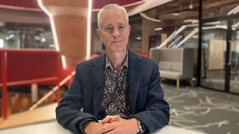 Jonathan Scourfield is wearing a patterned shirt and dark blazer. He is sitting in a modern office building.
Jonathan is looking straight at the camera. He has short spiky hair and blue eyes.