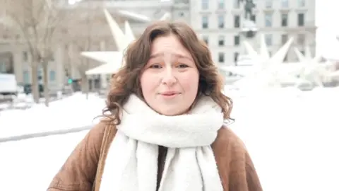 A young woman wearing a brown coat and white scarf speaking to the camera in a snowy square