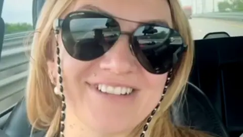 Polina Azarnykh looking straight at the camera while seated in a vehicle. She has long blonde hair and is wearing large sunglasses and a black top and is smiling.