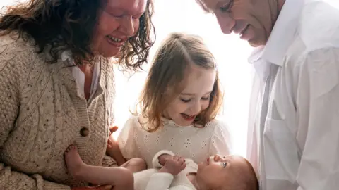 Aga Kowalska Photography A family portrait showing a mum dressed in an oatmeal cardigan and white shirt with a baby in a white baby grow, a girl in a white top and a man in a white shirt