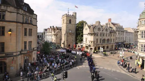 Oxford City Council With the sun shining on Oxford's medieval Carfax Tower, several dozen people have gathered behind temporary metal barriers to watch a ceremony.  A procession of people in suits can be seen and some choristers wearing traditional white dress 