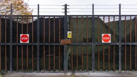 Barred gateway to the centre that has metal mesh between the gaps of the bars, barbed wire above it. On each gate there is a red octagonal stop sign. 