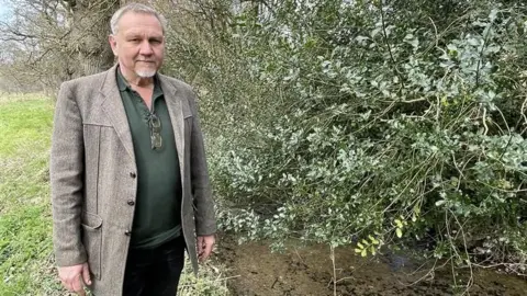 Alex Gater stood on the left next the water which has been contaminated by raw sewage. He is looking at the camer and is wearing a grey suit style jacket with a green shirt. The water has stones at the bottom and there is a large branch, with green leaves, hanging over the top of it.