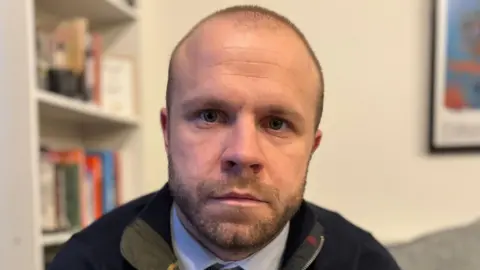 Man with short cropped hair and facial hair wearing a blue jumper and shirt