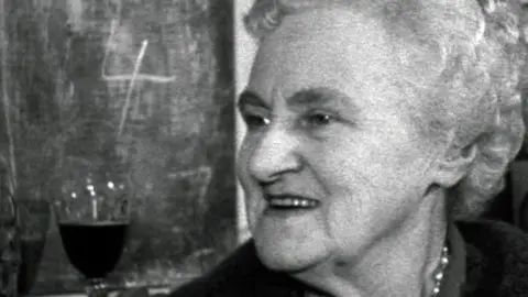 A woman smiling in a pub in 1962 with a glass of wine behind her.