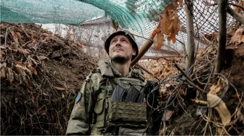 A Ukrainian soldier stands in a trench underneath a net of cover, looking up at the sky thoughtfully