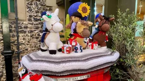 A knitted post box topper covering the top of a red post box. It features knitted children riding knitted horses with Christmas presents at the horses' hooves. The colourful topper is a tribute to the charity Gaddesden Place Riding for the Disabled. 