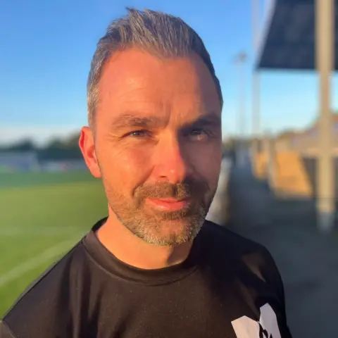 Close-up portrait of Gavin Roberts standing beside a football pitch in warm evening sunlight. Gavin is wearing a black sports top, with the background softly blurred to show part of a football field on the left.
Gavin has short grey hair with a spikey fringe. He also has a short, grey beard.