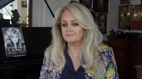 Bonnie Tyler sits in front of a piano with her autobiography on display