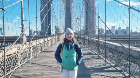 Paul Burke Becky Burke stood on Brooklyn Bridge in New York. The sun is shining, and New York's skyline can be seen in the background against a bright blue sky. Becky smiles at the camera with her hands in her pockets, she wears a navy and green raincoat, and has a tote bag over her shoulder.