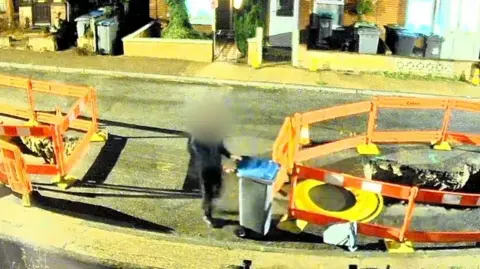 A person wearing black is on a residential road, with both hands on a black wheelie bin with a blue lid. Orange fencing encircles a hole in the road. It is nighttime.