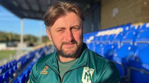 Callum Tipton sits in a football stadium stand wearing a dark green quarter-zip sports top with the “MENTRIBE FC” crest on the chest.
Callum has medium length brown hair with short sides and has a short beard.