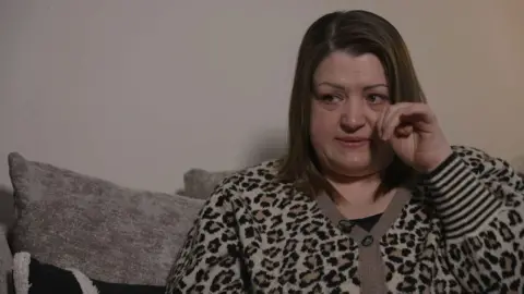 Gemma Robinson looking to the left of the camera. She has shoulder-length brown hair and brown eyes. She is wearing a beige leopard print knitted jumper and has her hand raised to her eye wiping away tears.