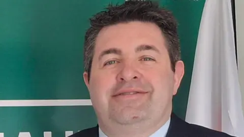 A man with short dark hair is smiling at the camera. He is wearing a dark suit and shirt. He is standing in front of a green and white background