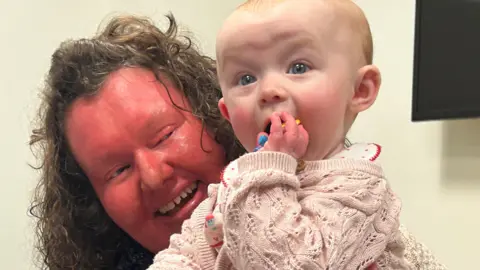 A woman with a red face and curly brown hair is holding a baby who is dressed in a pink jumper