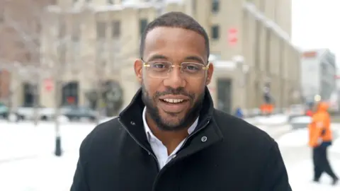 Man wearing glasses stands in front of buildings with snow on pavement