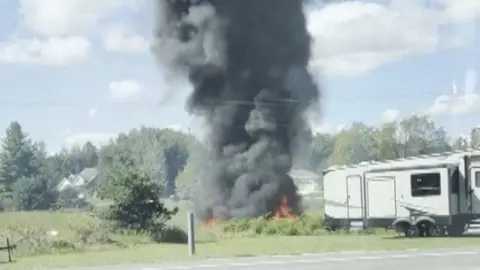 Flames and a plume of black smoke can be seen on a patch of grass near a caravan.