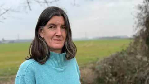 Rosie Pearson standing by a field with pylons in the background. She has shoulder-length brown hair and is wearing a light blue jumper.