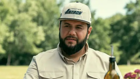 A still from the Netflix show Mo, showing the main character Mo wearing a cream coloured shirt and hat labelled "Houston Astros", as he gazes into the distance with the top of an olive oil bottle in front of him.