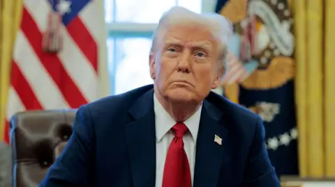 Donald Trump is wearing a navy suit jacket with a US flag pin, a white collared shirt and a red tie. He is looking up with a frown as he sits in the Oval Office, which has two flags on either side of him.