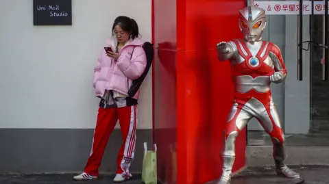 A young woman stands beside a life-sized Ultraman statue outside a shop.