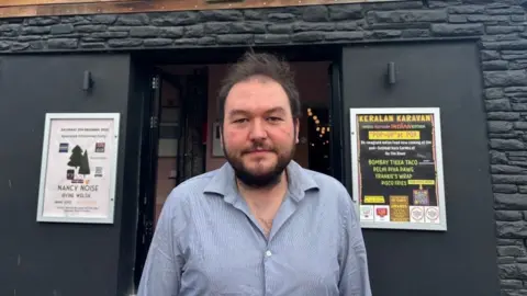 A man with black hair wearing a blue shirt stood outside a pub.