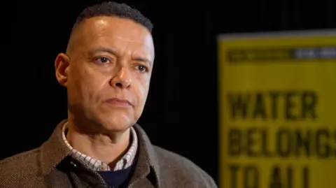 Cliver Lewis is left of the shot, standing inside and wearing a checked shirt, jumper and tweed jacket. He has short dark hair and there is a yellow sign in the background.