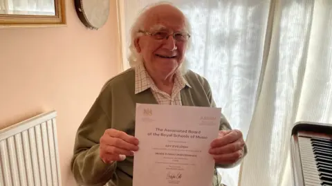 Ray Eveleigh, who has white hair and wears glasses and a green cardigan, holds up a certificate for passing his grade eight piano exam, while sitting next to his piano.