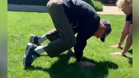 An elderly man has his hands on grass with bent knees facing the ground in a yoga position