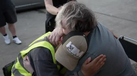 Mitchell hugs Peter Smorthit following his trek. Mitchell sits on a chair while Peter is in his wheelchair.