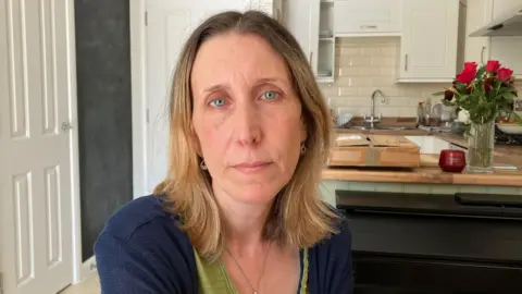 Lisa Lailey looking at the camera in front of her kitchen. Lisa has blonde hair and blue eyes. She is wearing a green top with a navy cardigan. 